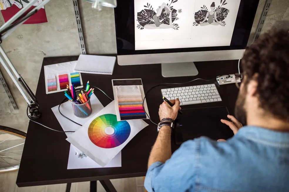 Designer gráfico trabalhando com mesa digitalizadora em estação de trabalho organizada com círculo cromático e paletas de cores.