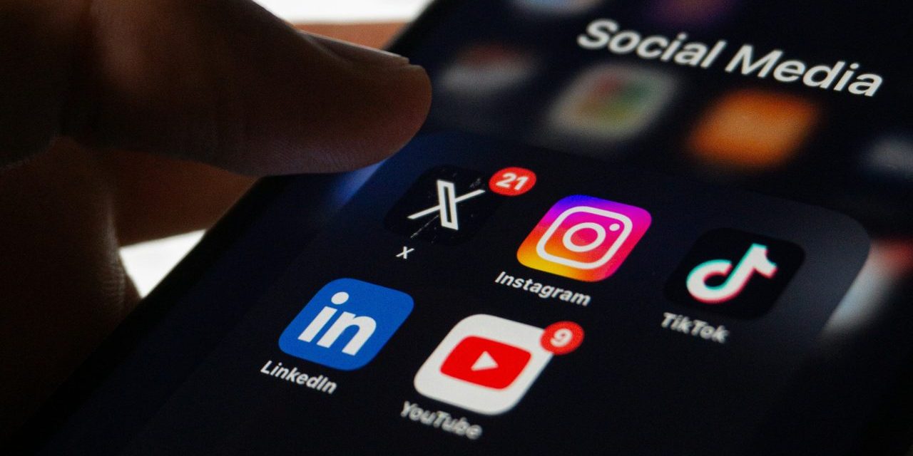 Pessoa segurando um celular no meio da cidade, com ícones de social media como redes sociais acima do aparelho.