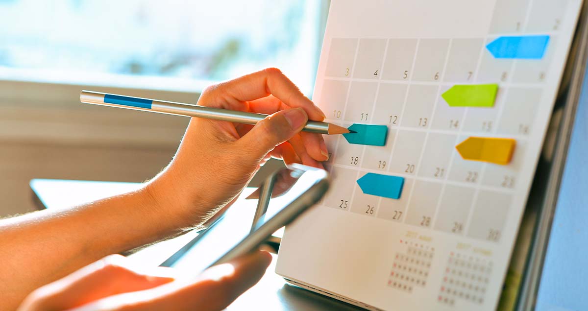 Uma mão segurando um lápis e apontando para um adesivo colorido em um calendário de mesa, com outra mão segurando um tablet ou smartphone. A imagem sugere organização e planejamento para estudar melhor.