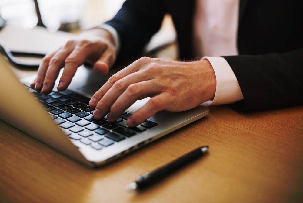 Uma pessoa com camisa e blazer, digitando em um laptop prateado sobre uma mesa de madeira. Uma caneta preta repousa ao lado do laptop. A imagem foca na ação de digitar, representando o engajamento com materiais de estudo ou pesquisa online para estudar melhor.