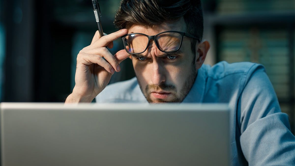 Uma visão em close-up de um homem jovem, com óculos pretos e camisa azul, olhando intensamente para a tela de um laptop, com uma expressão de concentração e uma caneta apoiada na têmpora. A imagem transmite a dedicação e o foco necessários para estudar melhor.