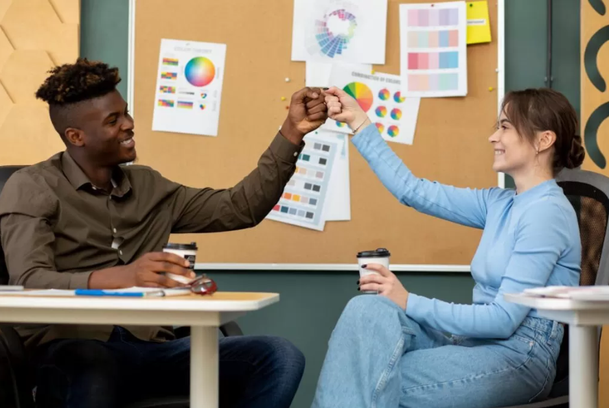 Uma imagem mostra duas pessoas, um homem e uma mulher, sorrindo e fazendo um "soquinho" (fist bump) com as mãos enquanto seguram copos de café. Eles estão sentados em uma mesa, com um quadro de avisos atrás deles cheio de gráficos de cores e amostras. A imagem representa o estudo colaborativo e o apoio mútuo como formas de estudar melhor.