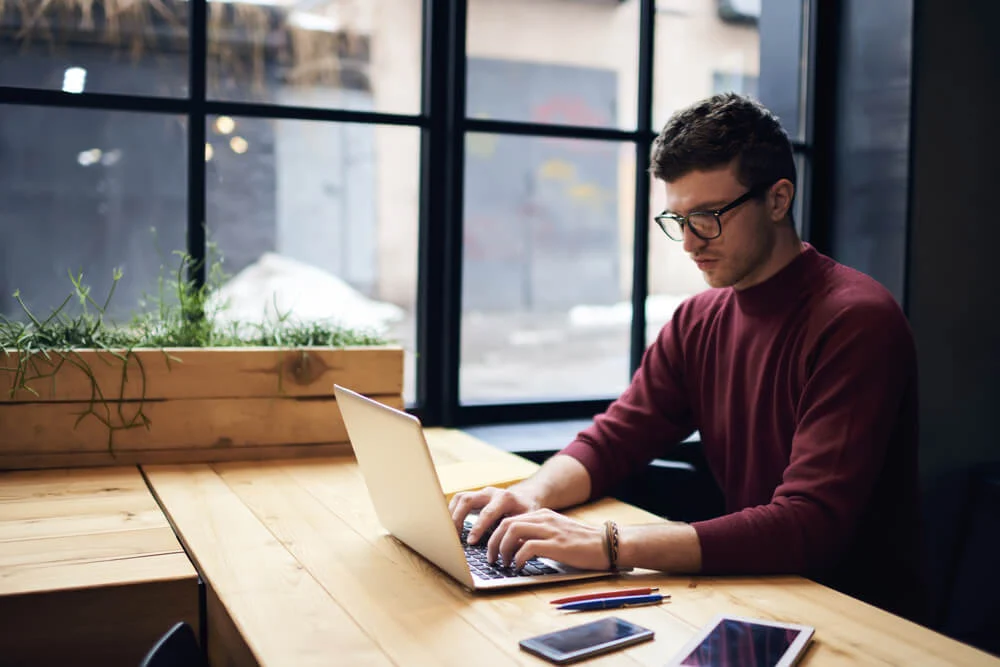 Homem concentrado trabalhando com copywriting em seu laptop, criando textos persuasivos em um ambiente moderno e inspirador, com dispositivos e materiais de apoio sobre a mesa.