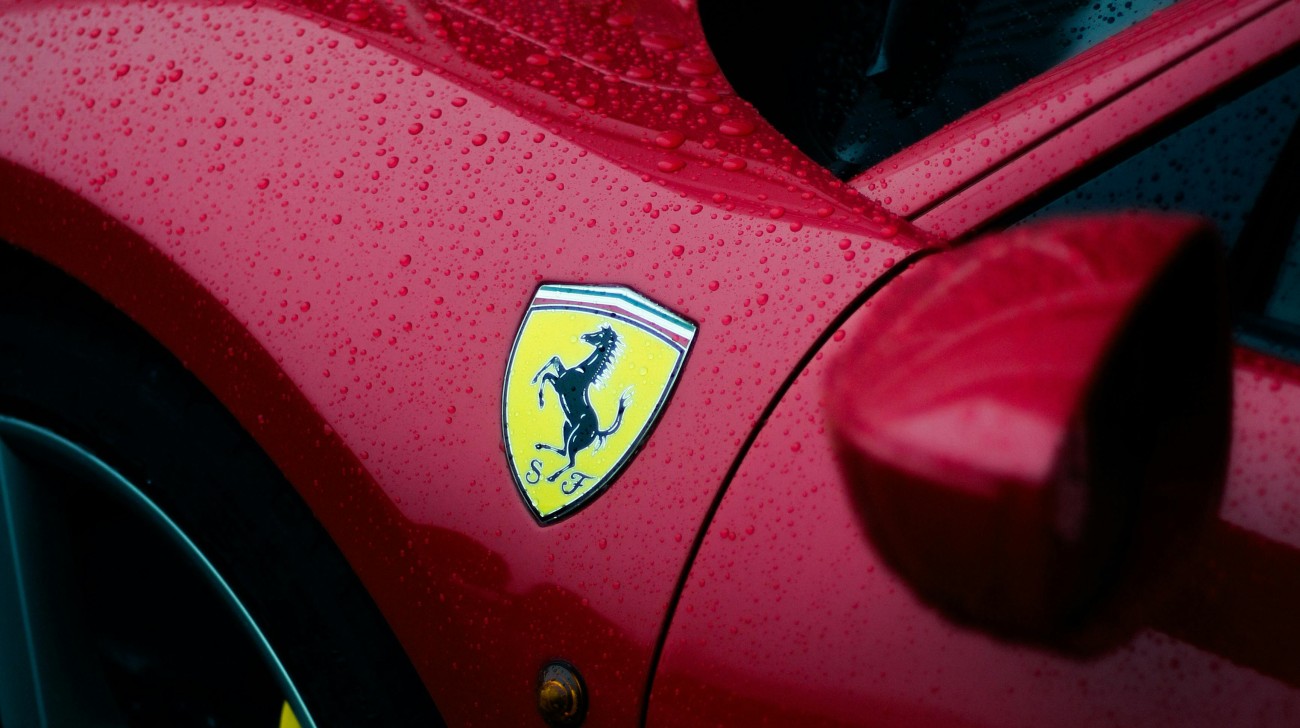 Uma imagem aproximada do escudo da Ferrari em um carro vermelho brilhante, coberto por gotas de chuva. O escudo amarelo vibrante exibe o cavalo empinado preto, que é um símbolo globalmente reconhecido de poder, velocidade e luxo, exemplificando os arquétipos do Herói e do Governante no branding automotivo.