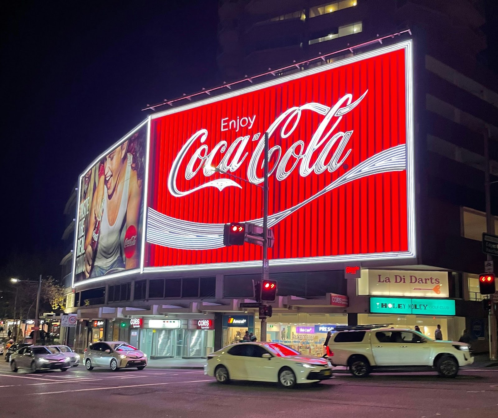 Um luminoso gigante da Coca-Cola, vermelho e brilhante, se destaca em uma rua movimentada à noite, com carros passando e edifícios ao fundo. A imagem de uma mulher sorrindo, bebendo Coca-Cola, aparece no lado esquerdo do luminoso, reforçando a associação da marca com a felicidade e o prazer, um exemplo clássico dos arquétipos da inocência e do prazer usados na publicidade.