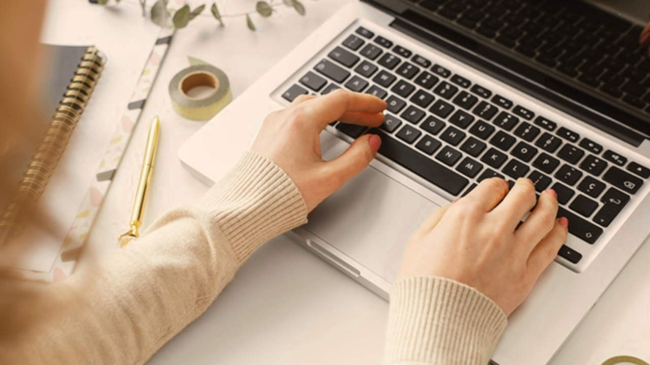 Uma visão aérea das mãos de uma pessoa digitando em um laptop, vestindo um suéter claro. Ao lado do laptop, há um caderno, uma caneta dourada e uma fita adesiva decorativa, sugerindo um ambiente de trabalho criativo ou estudo. A cena evoca o arquétipo do Sábio ou do Criador, representando a busca por conhecimento e a produção de ideias.