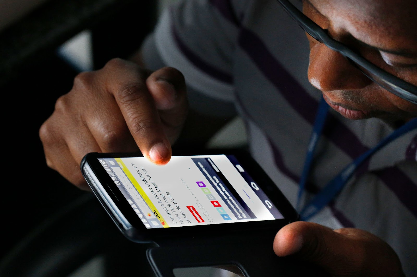 Um homem negro usando óculos examina atentamente o conteúdo de um smartphone. Seu dedo toca a tela, navegando por informações e representando a interação com o copywriting em dispositivos móveis.