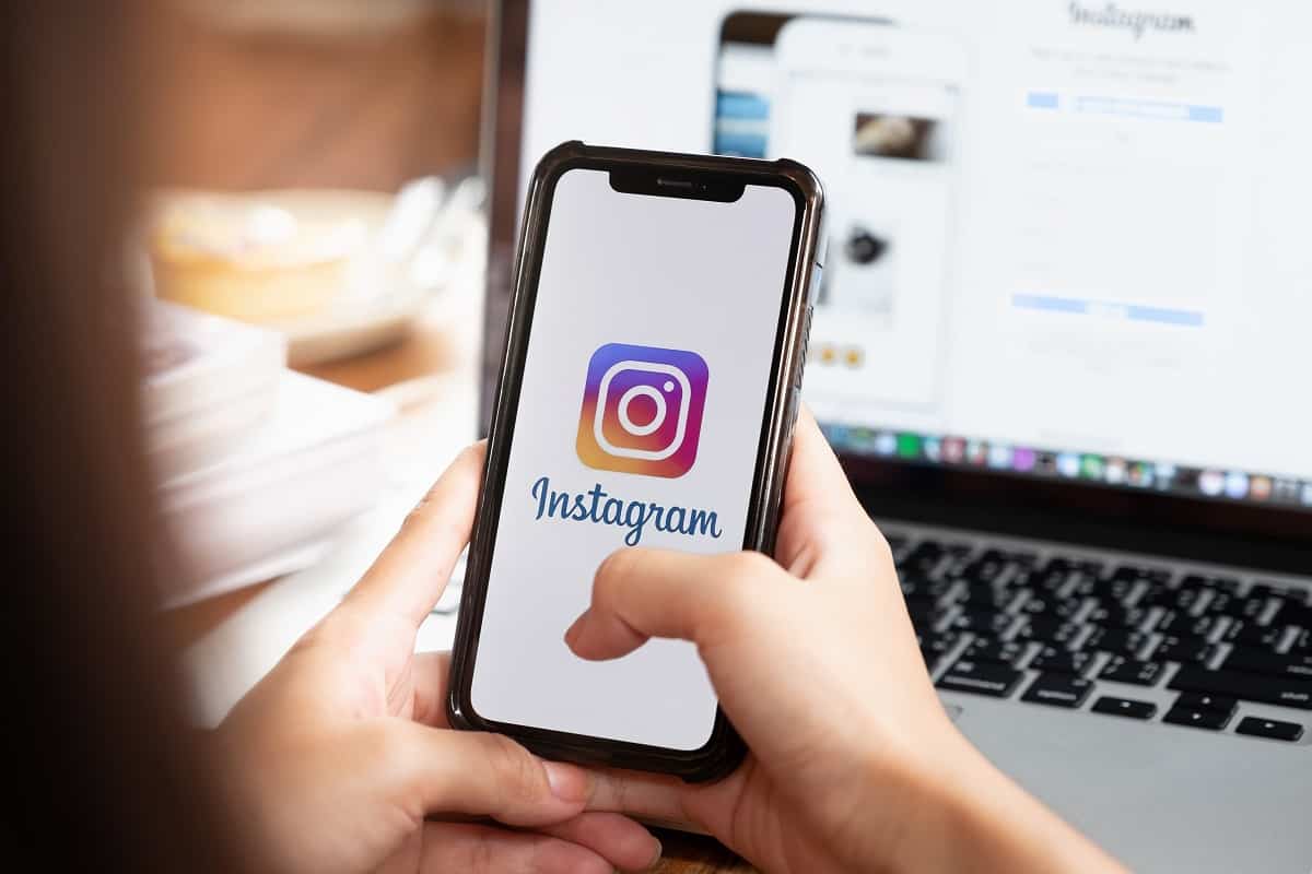 Uma pessoa segurando um smartphone com o aplicativo do Instagram aberto. A imagem mostra a tela do celular em primeiro plano, com um notebook ao fundo, simbolizando a gestão do "Alcance no Instagram" e estratégias de marketing digital.