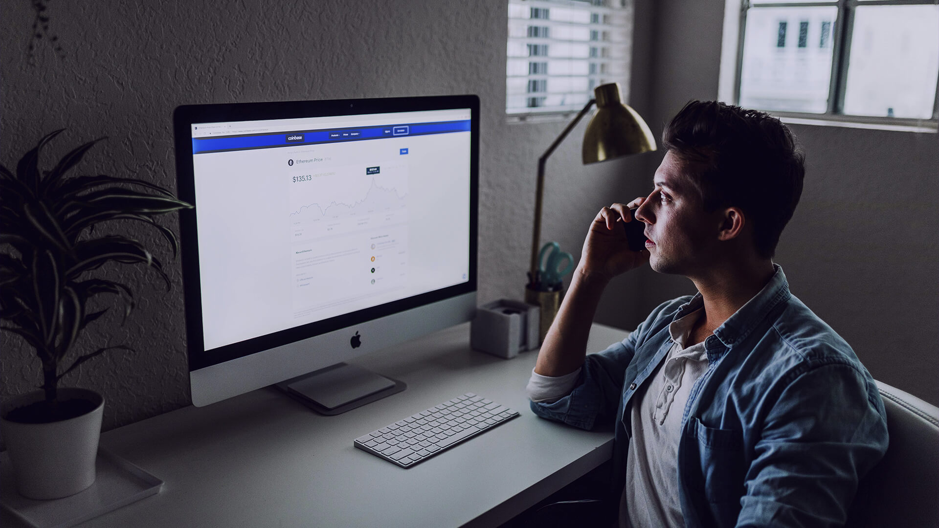 Um homem está sentado em uma mesa de escritório, olhando para a tela de seu computador que exibe um gráfico de preços de criptomoedas. Ele está segurando um celular ao lado da orelha, como se estivesse em uma ligação. A iluminação é suave e o ambiente parece ser um escritório em casa. A cena sugere a análise e o monitoramento de dados essenciais para o desenvolvimento de criativos de produtos digitais eficazes.