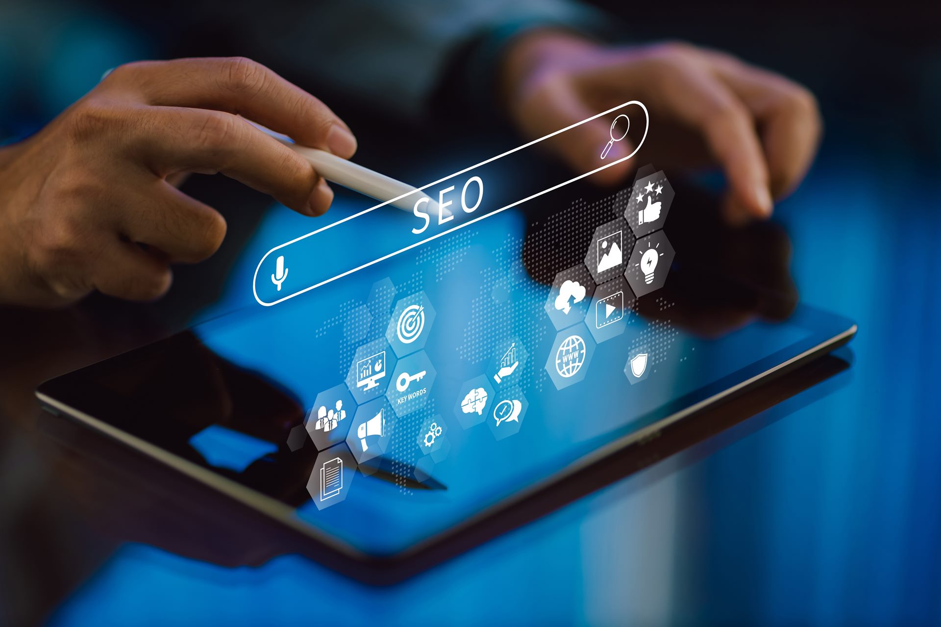 Um close-up mostra uma pessoa usando uma caneta digital para interagir com um tablet. Uma barra de pesquisa virtual com a palavra "SEO" está em destaque, e vários ícones hexagonais flutuam acima do dispositivo, simbolizando elementos de SEO Técnico, como a análise de dados, otimização de palavras-chave, e a usabilidade do site, tudo interligado por um mapa de rede digital.