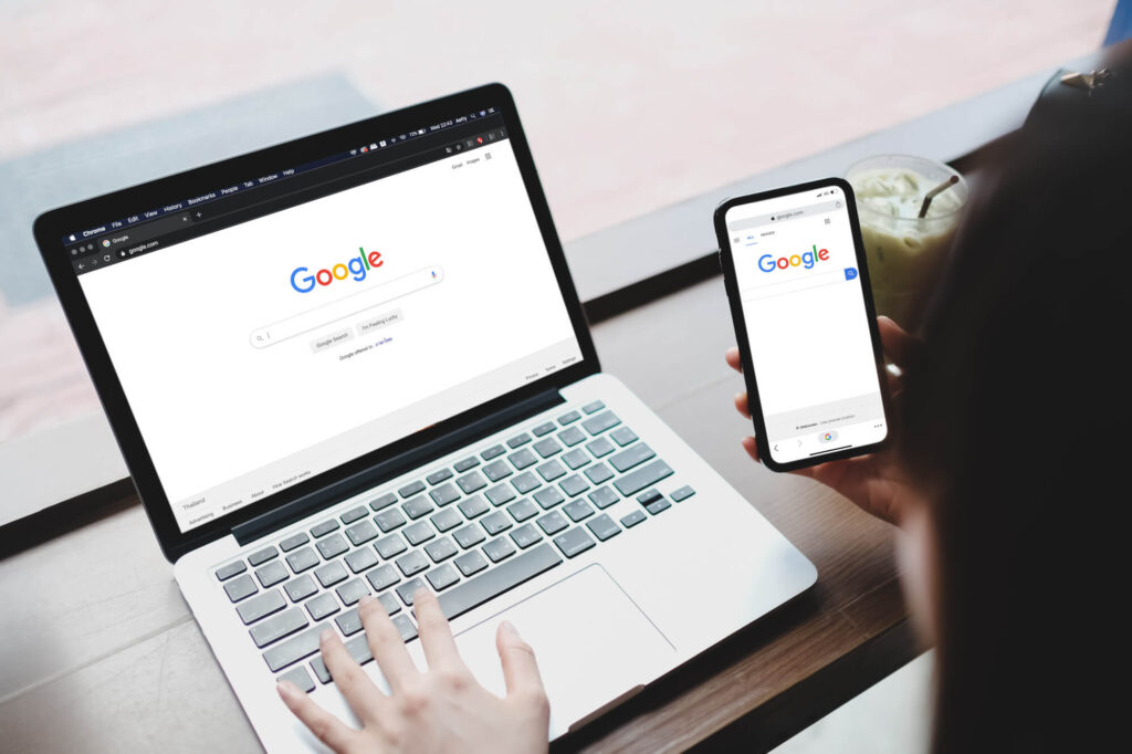 Uma pessoa está sentada em um café, com um notebook aberto na sua frente e um smartphone na mão. Ambos os dispositivos exibem a página inicial de pesquisa do Google. A imagem ilustra a importância de uma estratégia de SEO Técnico que considera a experiência do usuário em diferentes plataformas, como desktop e dispositivos móveis.
