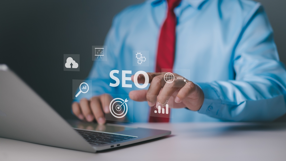 Um profissional em um escritório interage com ícones virtuais de SEO flutuando sobre um notebook. Sua mão aponta para a palavra "SEO", enquanto ícones como um alvo, um gráfico, uma nuvem e uma lupa representam os diferentes aspectos do SEO Técnico, incluindo a análise de dados, otimização de performance e a experiência do usuário.